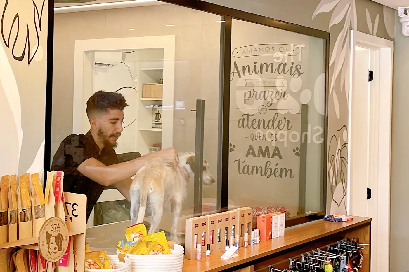 A sala de banho e tosa vista pelo vidro: um rapaz de camiseta preta com um cachorro claro na mesa e, no vidro, o adesivo com a frase da loja.